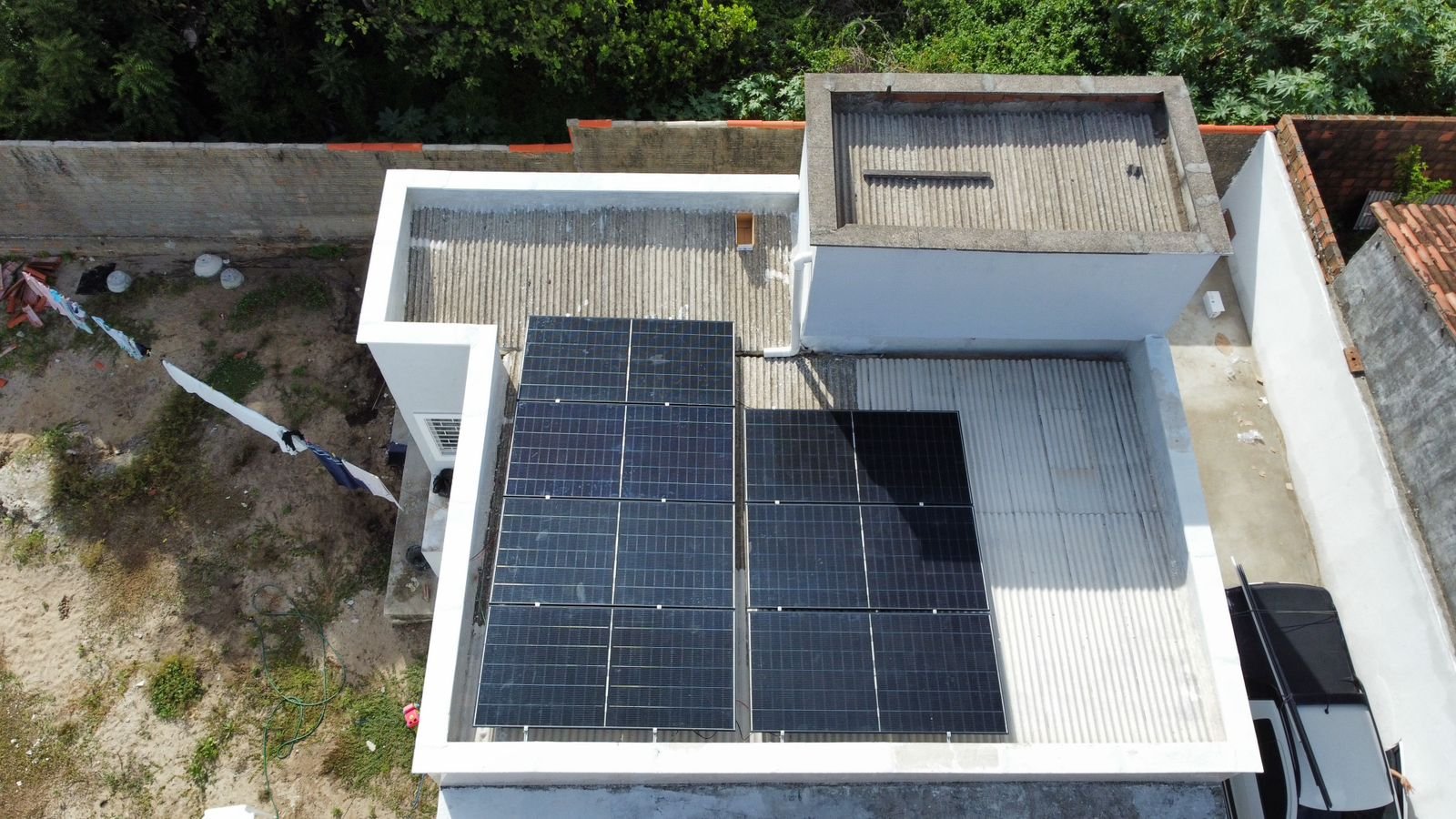 Vista aérea de telhado residencial com placas de energia solar instaladas ao lado de um condicionador de ar tipo split montado na parede, quintal de terra com roupas no varal ao fundo.