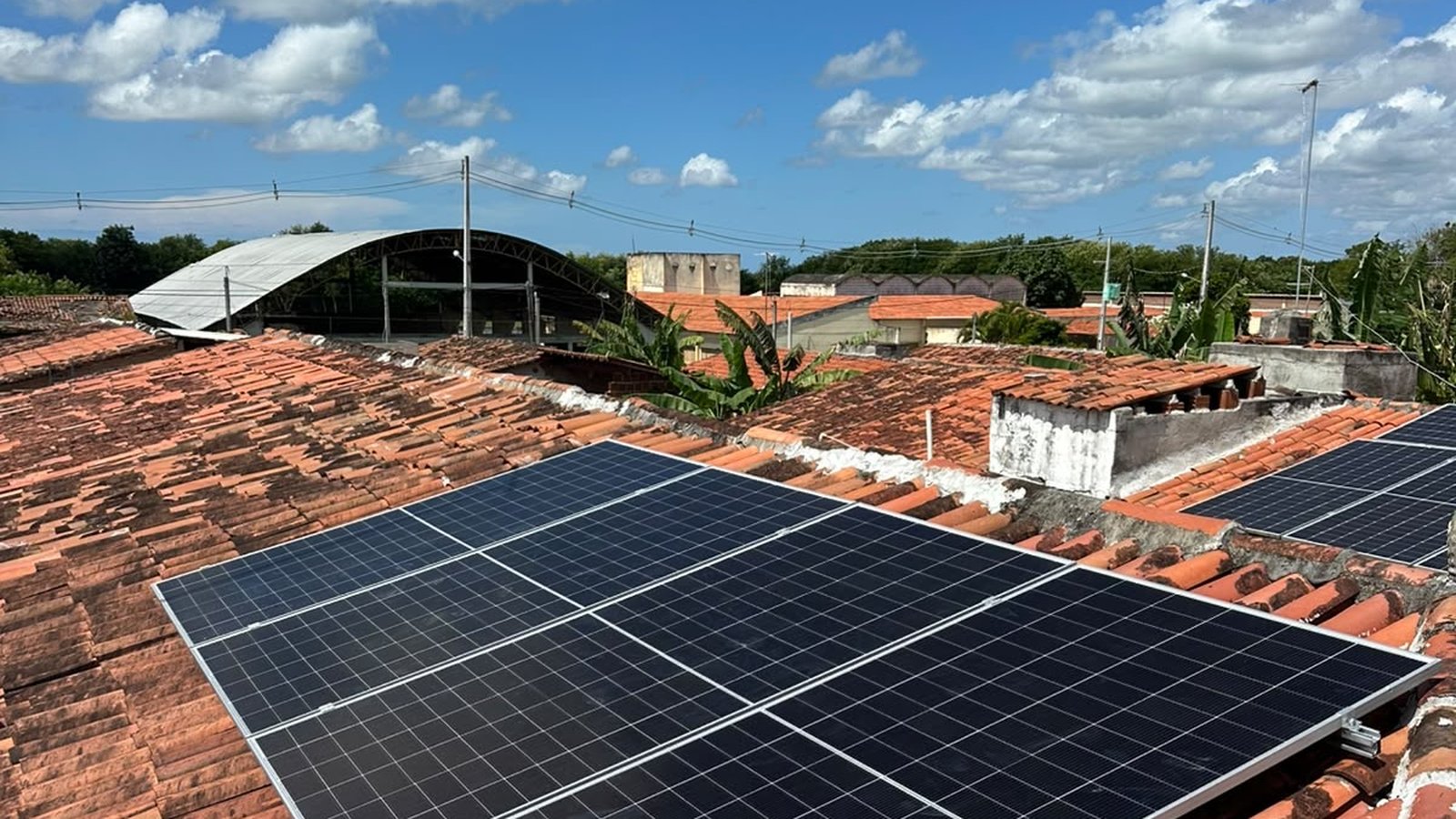 Placas de energia solar instaladas em telhado de telha cerâmica, fotografadas de baixo para cima contra céu azul de verão com nuvens brancas, em dia de sol forte.