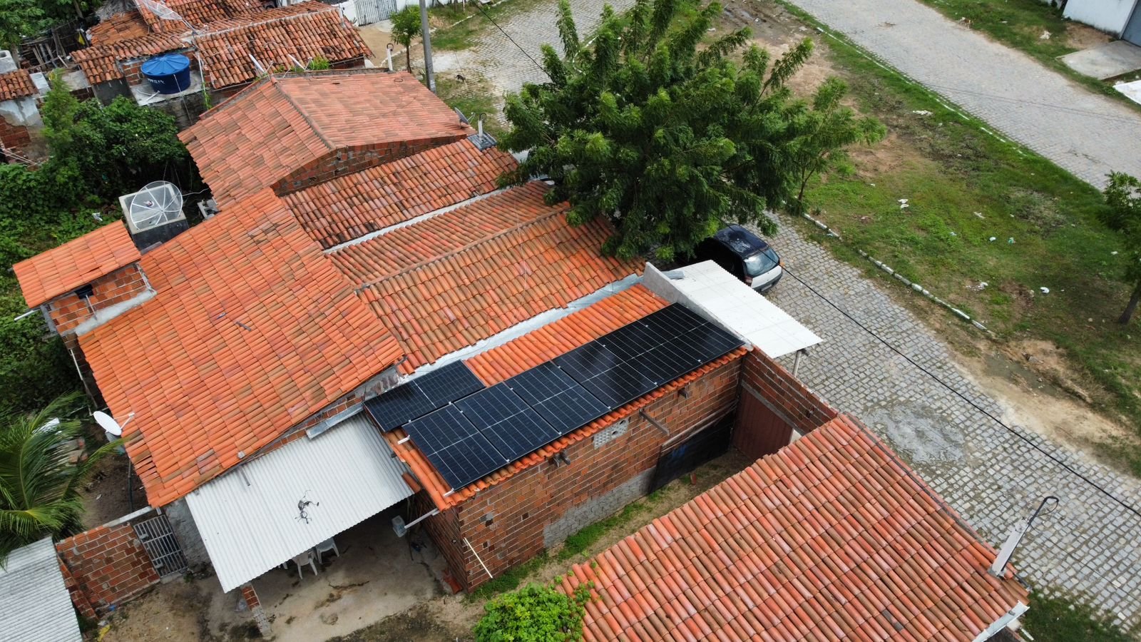 Vista de drone de um grupo de casas vizinhas de telha cerâmica, em que só uma tem módulos solares no telhado, com árvores ao redor.