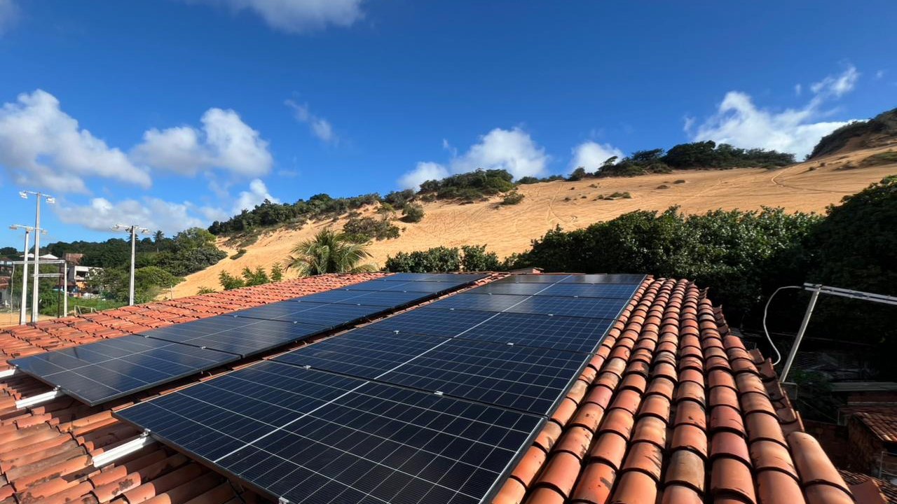 Módulos solares montados num telhado de telha cerâmica, com céu azul e dunas ao fundo.