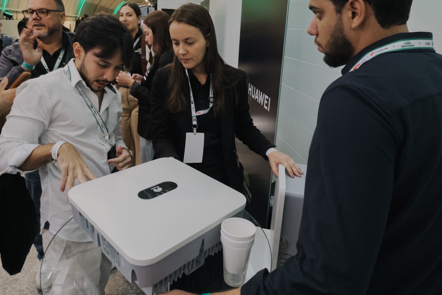 Três pessoas examinando um inversor Huawei em exposição no corredor de estandes do evento.
