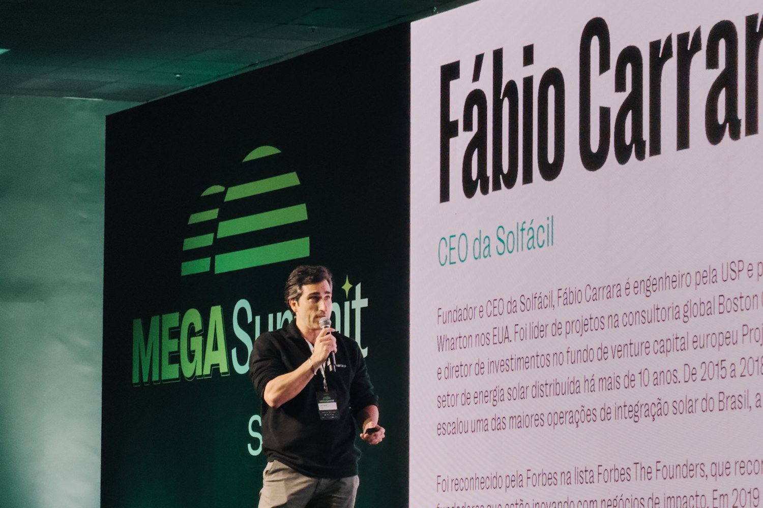Fábio Carrara no palco do Mega Summit, de microfone na mão, diante de um telão que exibe seu nome e o cargo de CEO da Solfácil.