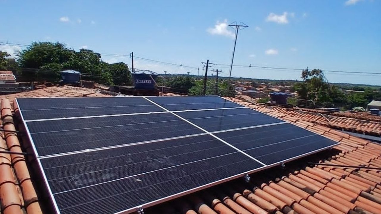 Usina solar grande sobre telhado de telha cerâmica, com duas fileiras longas de módulos fotovoltaicos, céu azul e postes de energia ao fundo.