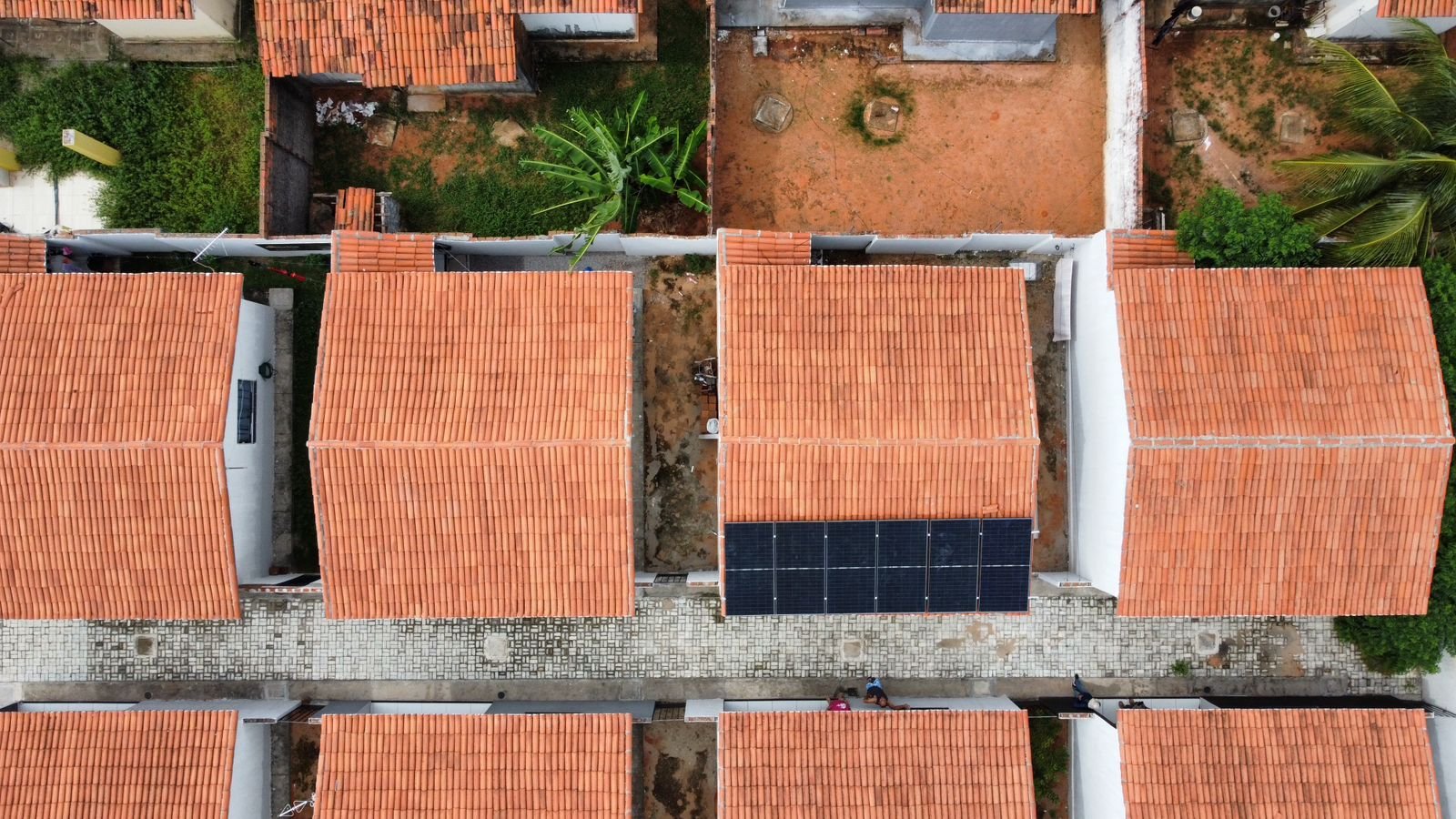 Vista de drone de uma casa pequena de telha cerâmica com poucos módulos solares no telhado, entre telhados vizinhos.