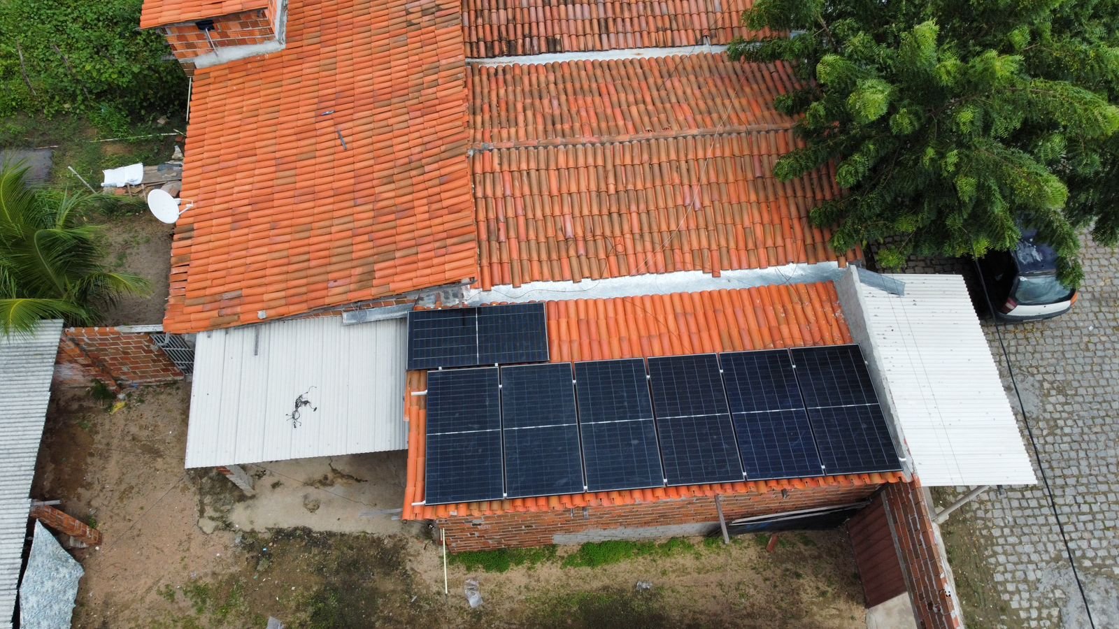 Vista aérea de casa residencial com usina de energia solar no telhado, cercada por árvores, com carro estacionado na garagem ao lado.