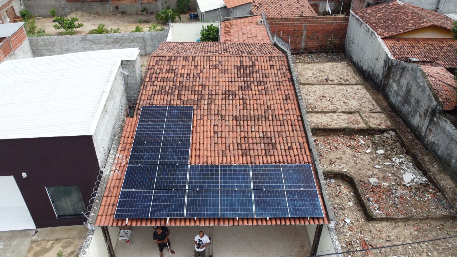 Vista de drone de uma casa com telhado cerâmico e 18 módulos solares instalados em dois blocos, com um terreno com entulho ao lado, em um bairro residencial.