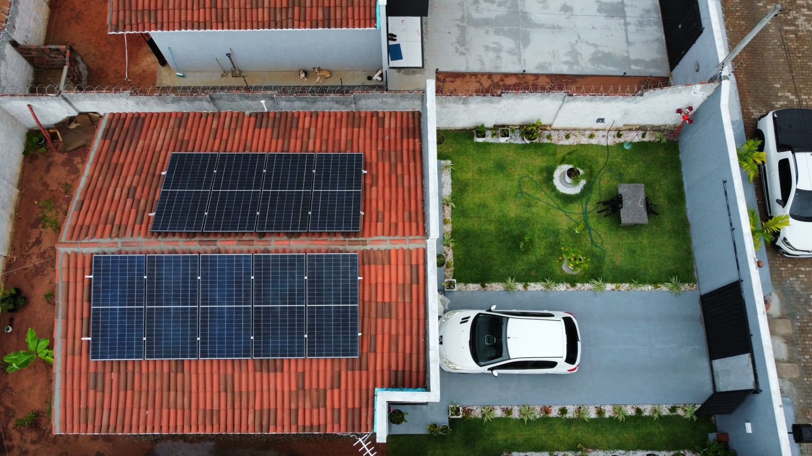 Vista de drone de uma casa com telhado cerâmico e módulos solares instalados em duas águas, com gramado e carro na garagem, na Grande Natal.