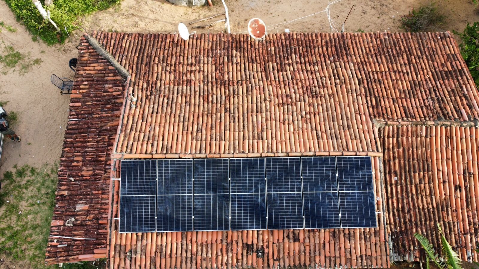 Vista aérea aproximada de sete módulos solares instalados em fileira sobre um telhado de telha cerâmica, com vegetação e uma rua ao fundo.