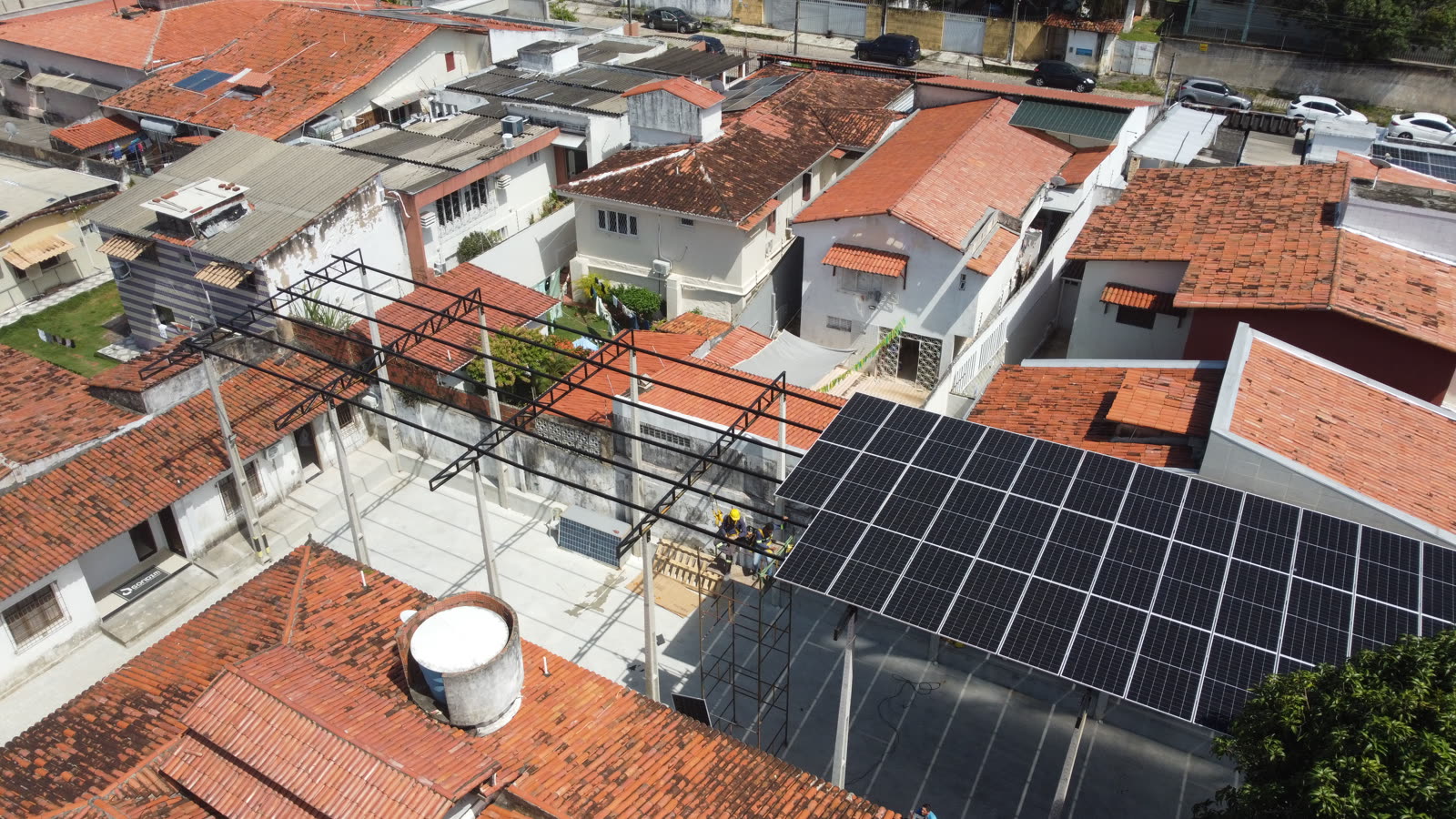 Vista aérea de uma cobertura solar em construção sobre o pátio de uma empresa em Natal, com módulos fotovoltaicos apoiados em estrutura metálica e telhados de telha cerâmica ao redor.