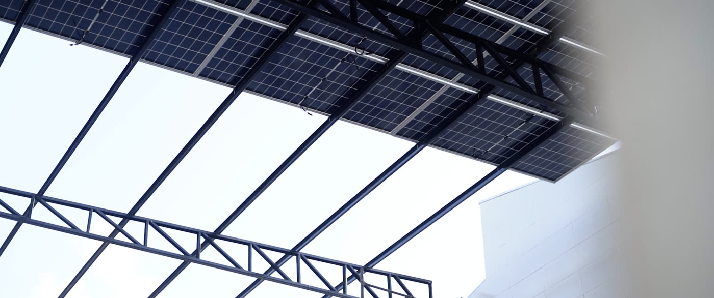 Vista de baixo da cobertura solar: os módulos fotovoltaicos apoiados sobre treliças metálicas formam o próprio teto do pátio, deixando vãos abertos para o céu.