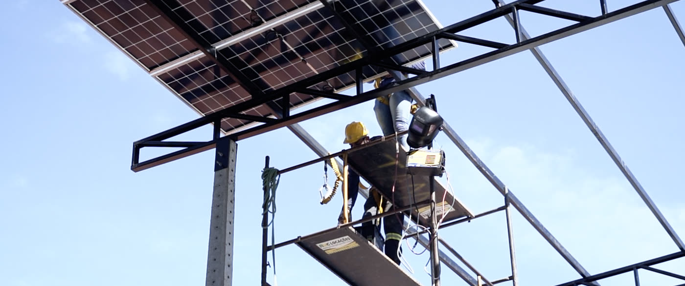 Instalador solda a estrutura metálica já com módulos fotovoltaicos instalados acima, trabalhando sobre andaime com máscara de solda ao lado.