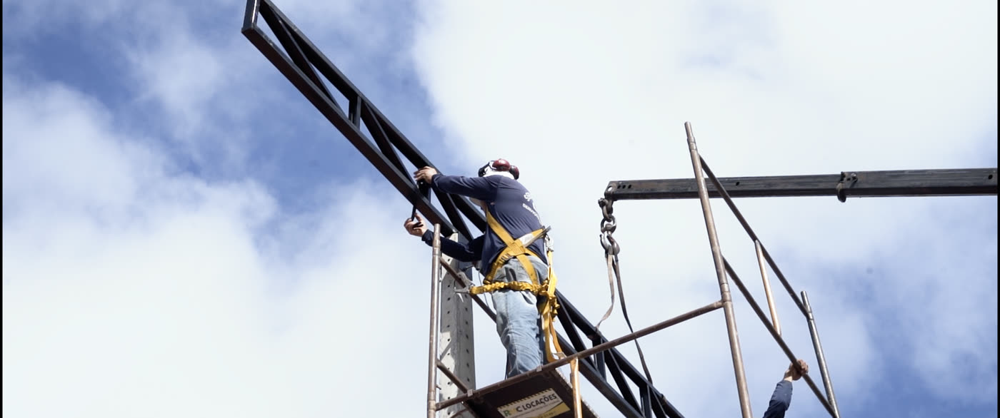 Instalador com cinto de segurança sobre andaime posiciona uma treliça metálica içada por corrente, encaixando-a no topo de um poste de concreto.
